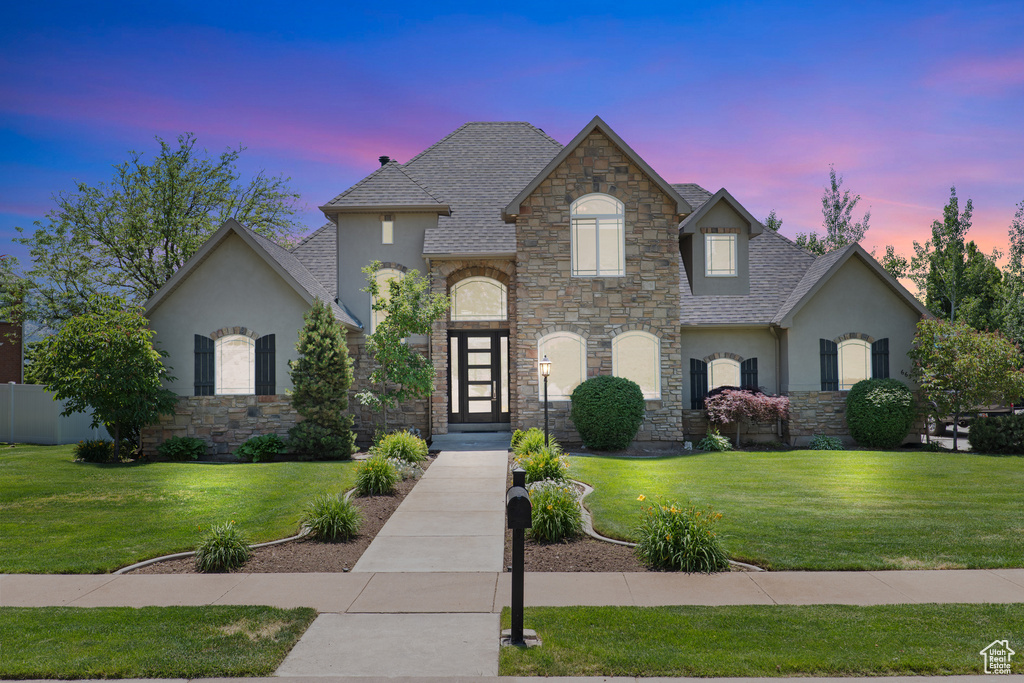 French country inspired facade with stucco siding, stone siding, a front lawn, and roof with shingles
