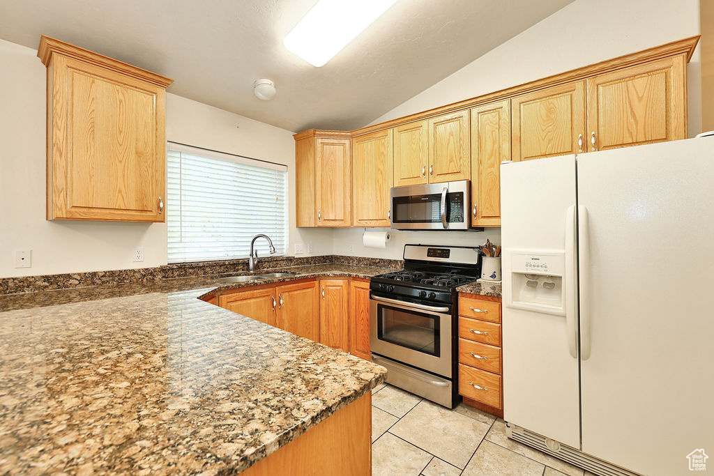 Kitchen with appliances with stainless steel finishes, vaulted ceiling, light tile patterned flooring, light brown cabinets, and dark stone counters