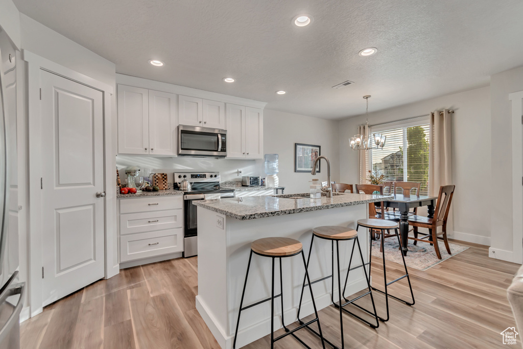 Kitchen with appliances with stainless steel finishes, light wood-style flooring, a chandelier, white cabinetry, and light stone countertops