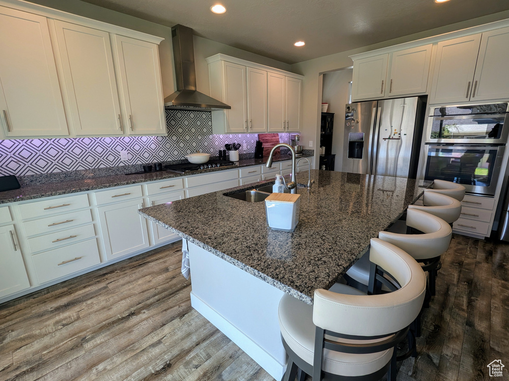 Kitchen featuring wall chimney exhaust hood, appliances with stainless steel finishes, white cabinetry, recessed lighting, and wood finished floors