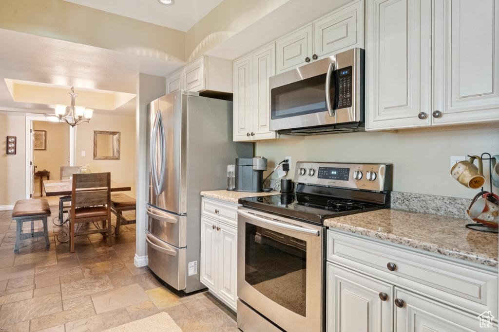 Kitchen with appliances with stainless steel finishes, a chandelier, white cabinetry, stone tile flooring, and light stone countertops