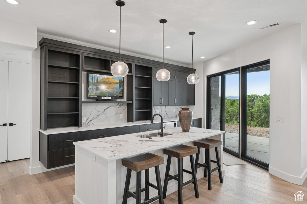 Kitchen featuring open shelves, a center island with sink, tasteful backsplash, light wood-type flooring, and pendant lighting