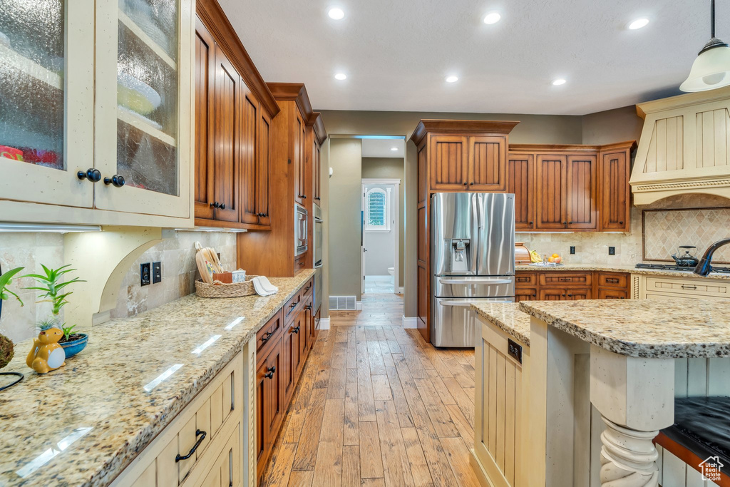 Kitchen featuring appliances with stainless steel finishes, tasteful backsplash, premium range hood, glass insert cabinets, and brown cabinets