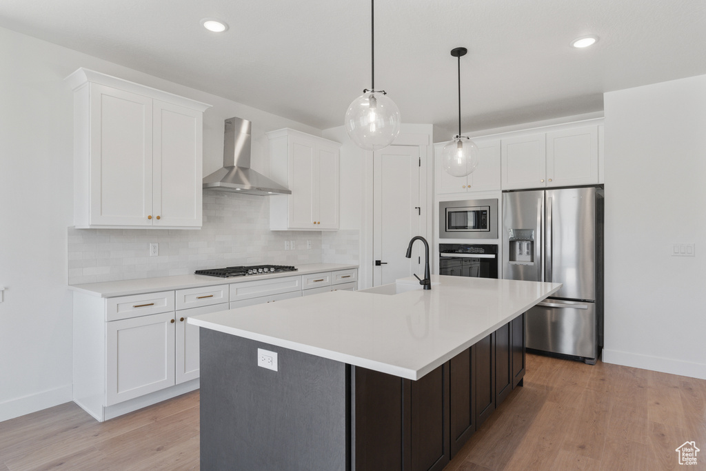 Kitchen featuring wall chimney range hood, appliances with stainless steel finishes, decorative backsplash, light countertops, and a center island with sink