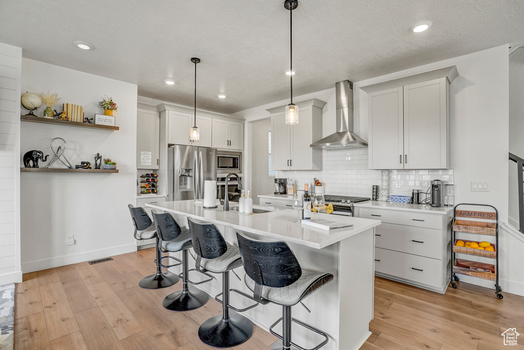 Kitchen featuring wall chimney range hood, appliances with stainless steel finishes, a kitchen breakfast bar, backsplash, and light countertops