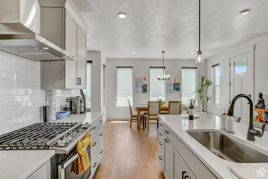 Kitchen with wall chimney exhaust hood, stainless steel gas range, pendant lighting, tasteful backsplash, and light stone countertops