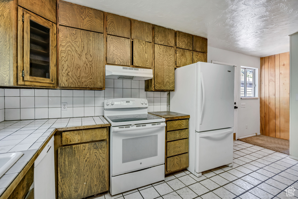 Kitchen with under cabinet range hood, white appliances, tile counters, backsplash, and light tile patterned floors