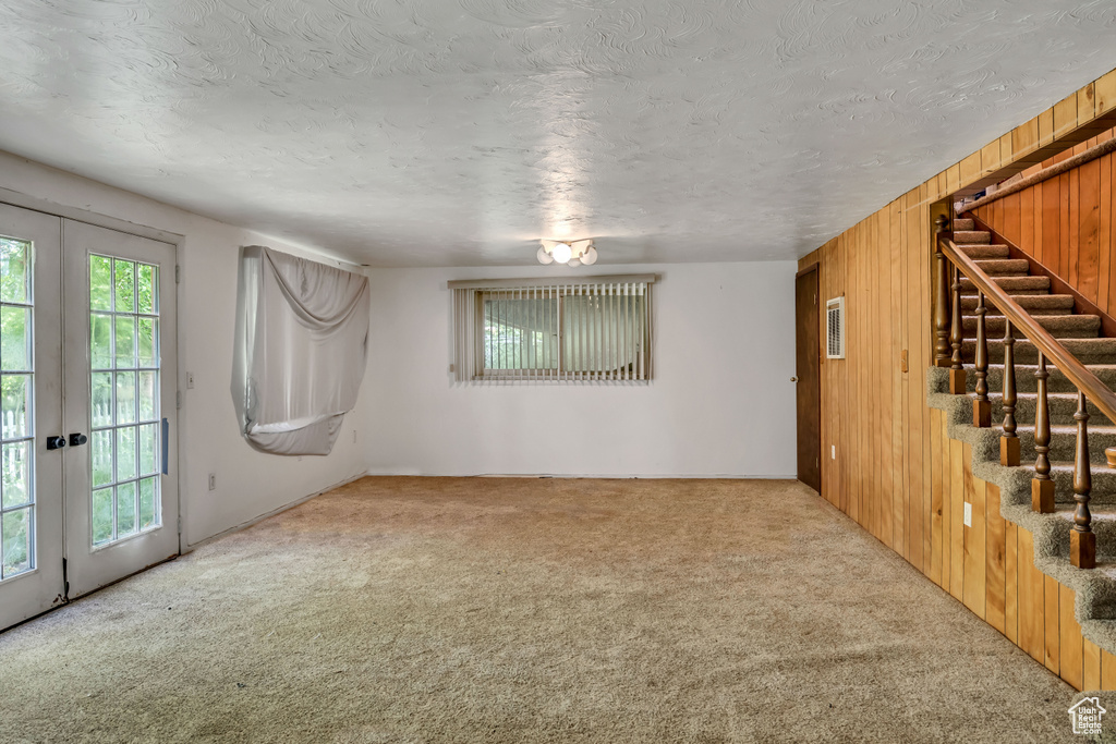 Unfurnished living room featuring carpet flooring, french doors, stairway, wood walls, and a textured ceiling