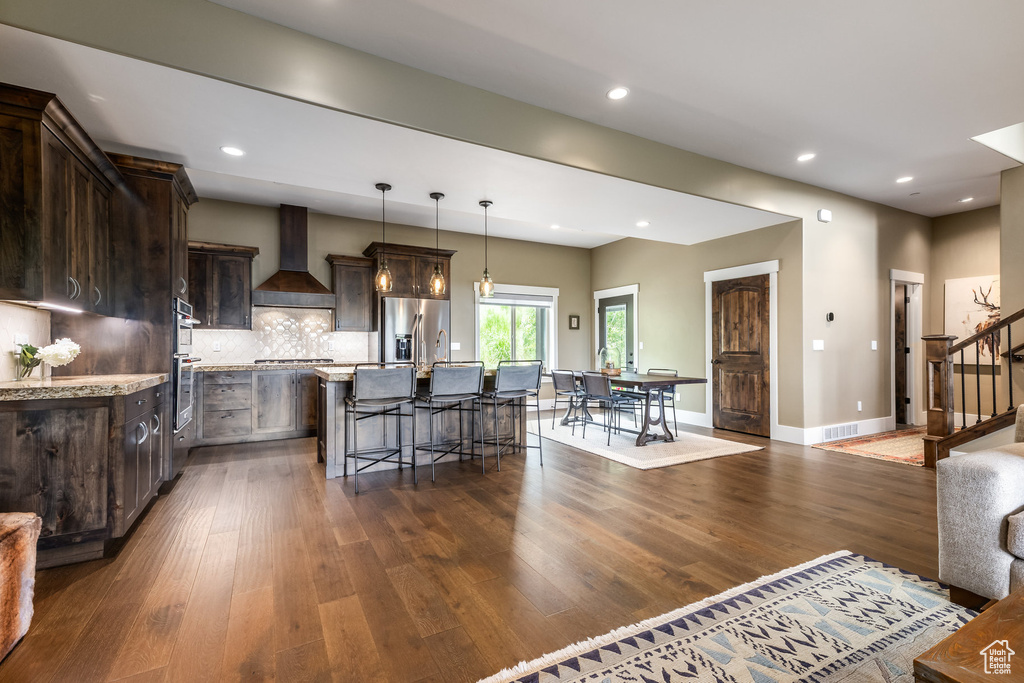 Kitchen featuring wall chimney range hood, stainless steel refrigerator with ice dispenser, dark brown cabinetry, an island with sink, and dark wood finished floors