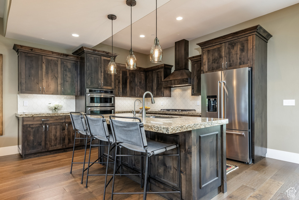 Kitchen featuring stainless steel appliances, custom exhaust hood, wood finished floors, dark brown cabinets, and recessed lighting