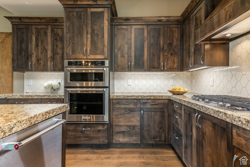 Kitchen featuring stainless steel appliances, wall chimney range hood, dark brown cabinets, decorative backsplash, and dark wood-style floors