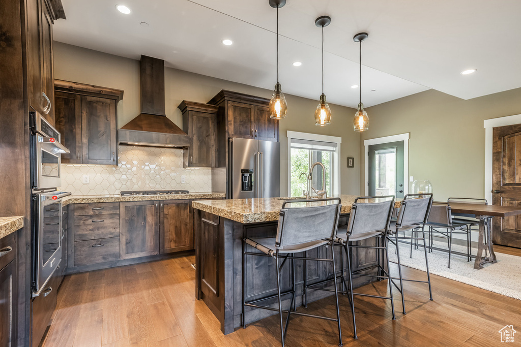 Kitchen with stainless steel appliances, wall chimney range hood, wood finished floors, dark brown cabinets, and recessed lighting