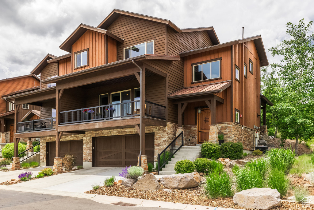 View of front of property featuring stone siding, a balcony, board and batten siding, an attached garage, and a standing seam roof