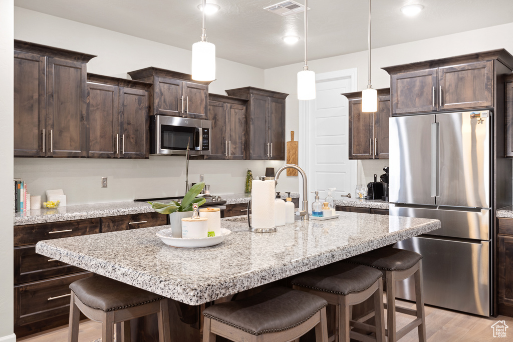 Kitchen with appliances with stainless steel finishes, a kitchen breakfast bar, dark brown cabinets, light wood finished floors, and an island with sink