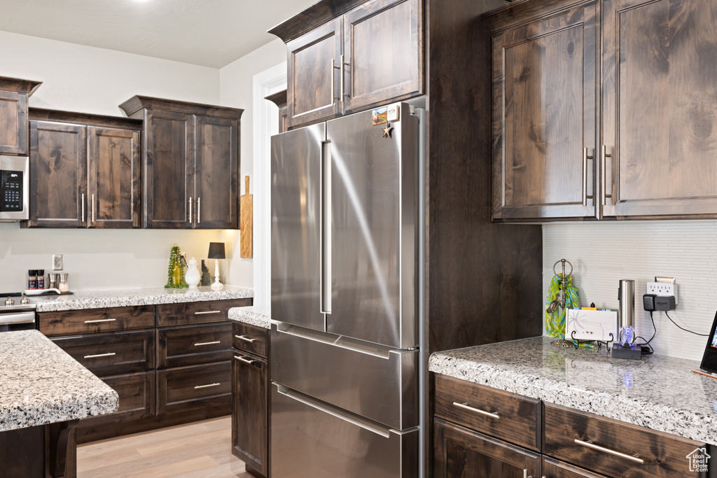 Kitchen with appliances with stainless steel finishes, dark brown cabinets, light stone countertops, and light wood-type flooring