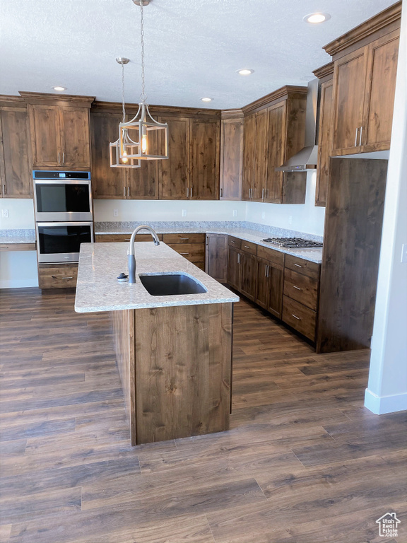 Kitchen featuring wall chimney exhaust hood, appliances with stainless steel finishes, recessed lighting, hanging light fixtures, and an island with sink
