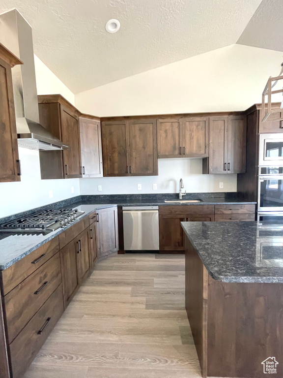 Kitchen featuring appliances with stainless steel finishes, vaulted ceiling, wall chimney range hood, light wood-type flooring, and a textured ceiling