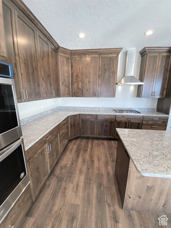 Kitchen featuring wall chimney range hood, stainless steel appliances, dark wood-style flooring, a textured ceiling, and recessed lighting