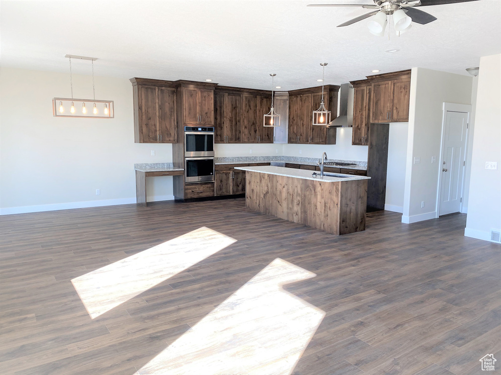 Kitchen with wall chimney exhaust hood, double oven, ceiling fan, a kitchen island with sink, and dark brown cabinetry