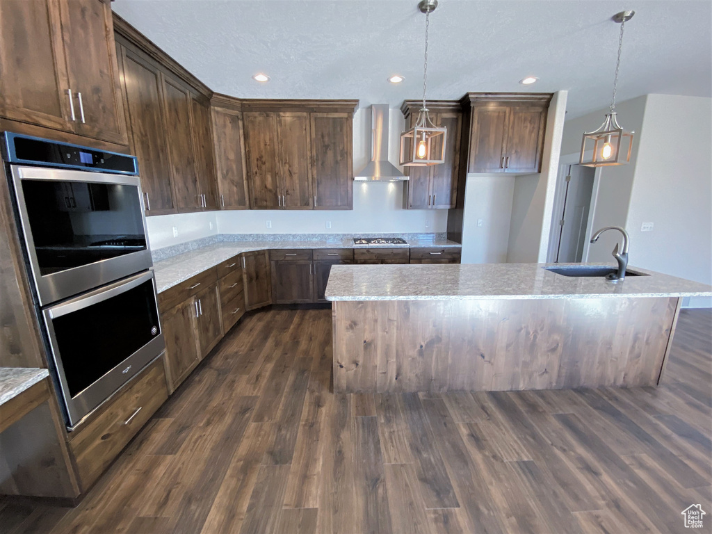 Kitchen with stainless steel appliances, wall chimney range hood, dark wood finished floors, a center island with sink, and pendant lighting