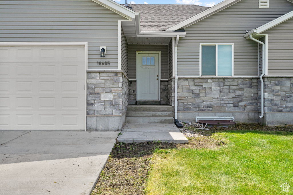 View of exterior entry with stone siding, an attached garage, a yard, driveway, and roof with shingles