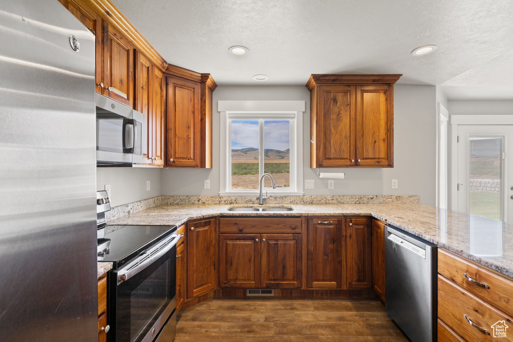 Kitchen with stainless steel appliances, dark wood-type flooring, brown cabinets, light stone countertops, and recessed lighting