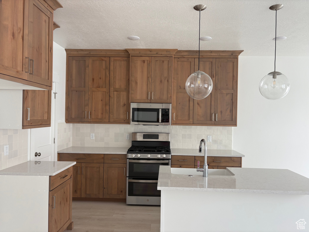 Kitchen featuring appliances with stainless steel finishes, light wood-style flooring, brown cabinetry, decorative backsplash, and a textured ceiling