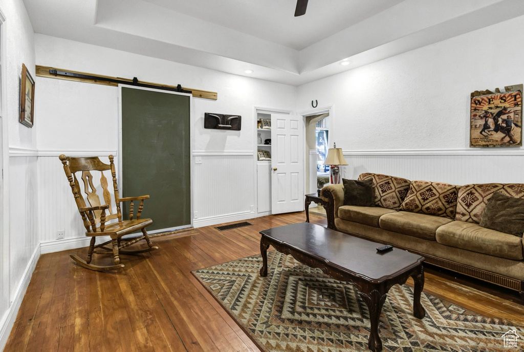Living room featuring a barn door, wood-type flooring, a tray ceiling, ceiling fan, and a wainscoted wall