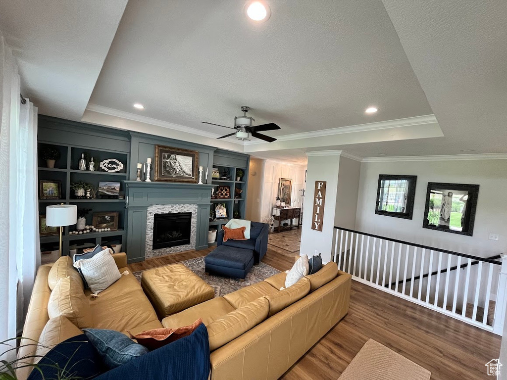 Living area featuring a tray ceiling, wood finished floors, a tiled fireplace, a ceiling fan, and crown molding