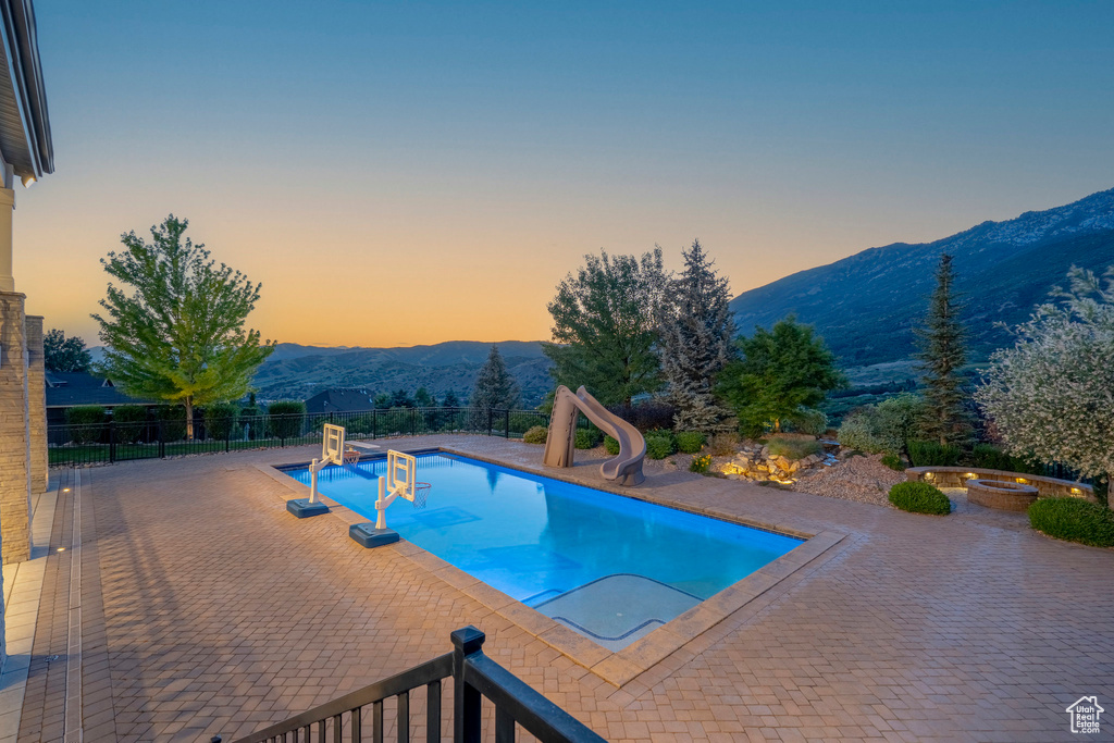 Pool at dusk featuring a water slide, a mountain view, a hot tub, and a patio