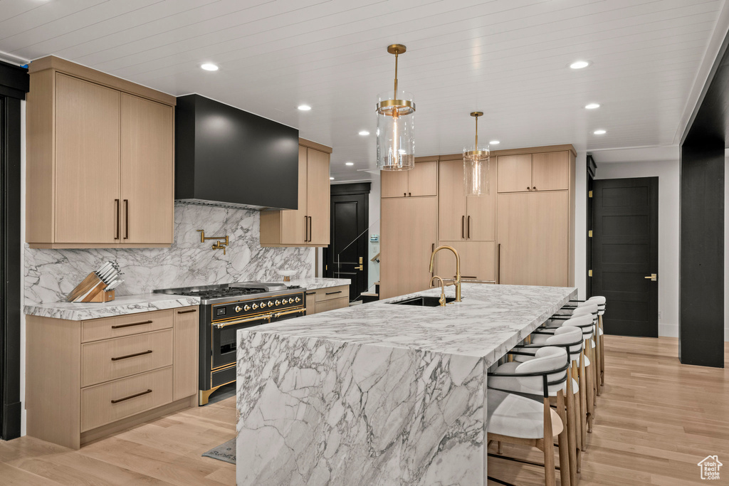 Kitchen with stainless steel stove, light brown cabinetry, range hood, backsplash, and light wood-style floors