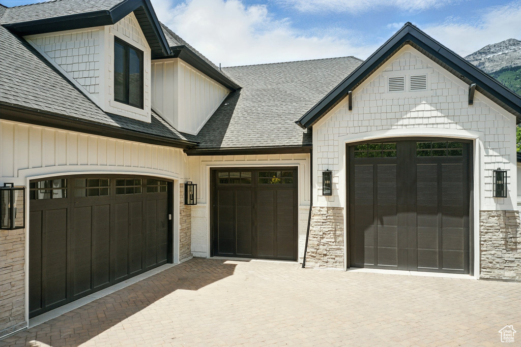 View of front facade with roof with shingles, decorative driveway, stone siding, and an attached garage