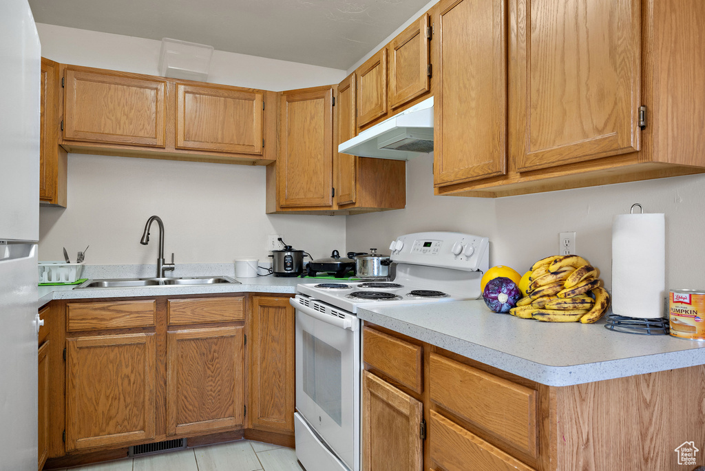 Kitchen with white range with electric stovetop, under cabinet range hood, freestanding refrigerator, light countertops, and brown cabinetry