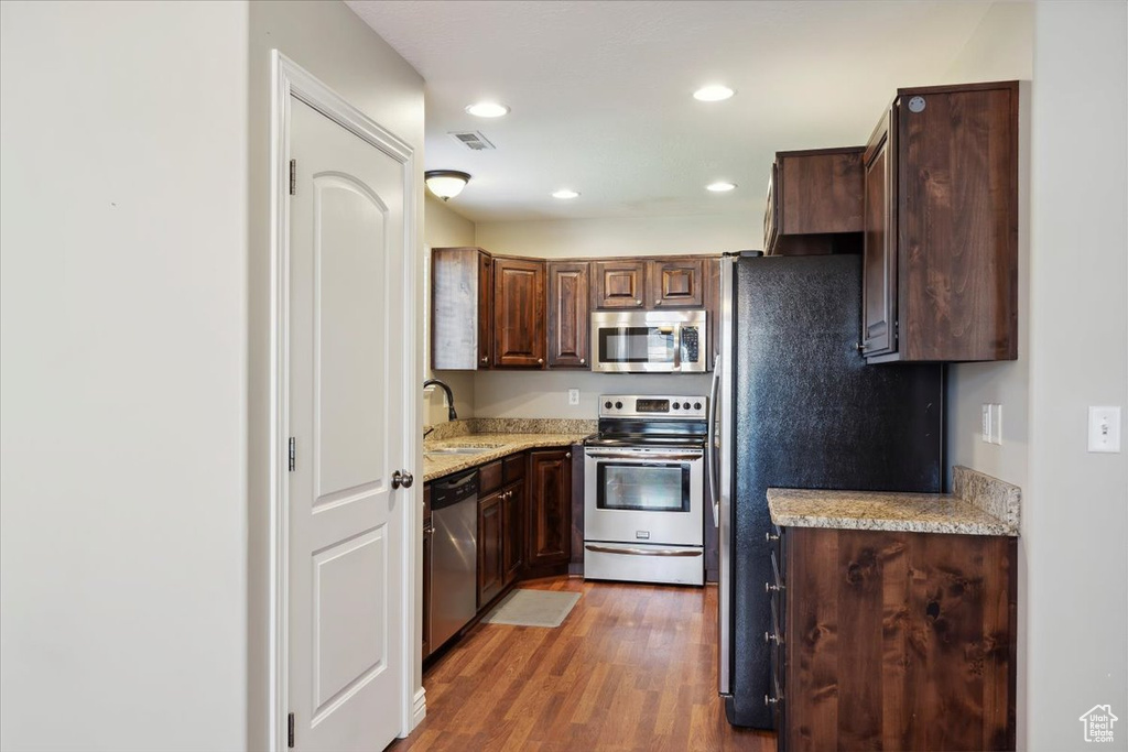 Kitchen featuring appliances with stainless steel finishes, wood finished floors, recessed lighting, dark brown cabinetry, and light stone countertops