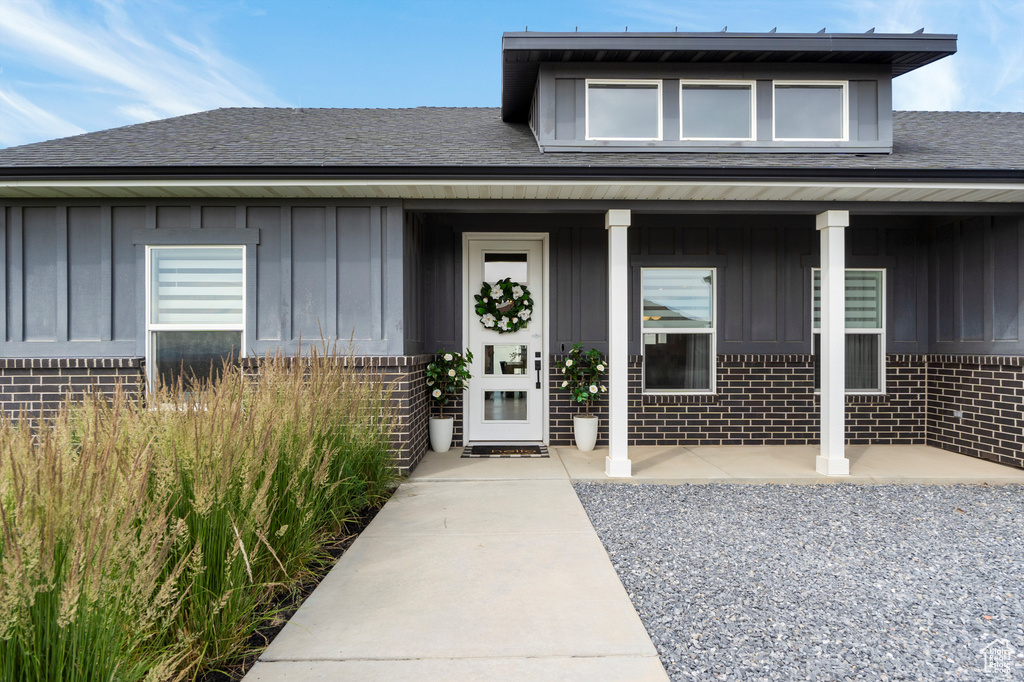 View of exterior entry with board and batten siding, brick siding, a shingled roof, and a porch