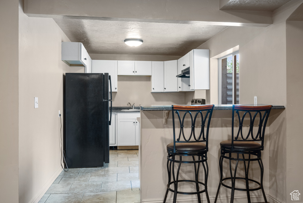 Kitchen featuring freestanding refrigerator, a breakfast bar, dark countertops, under cabinet range hood, and white cabinetry