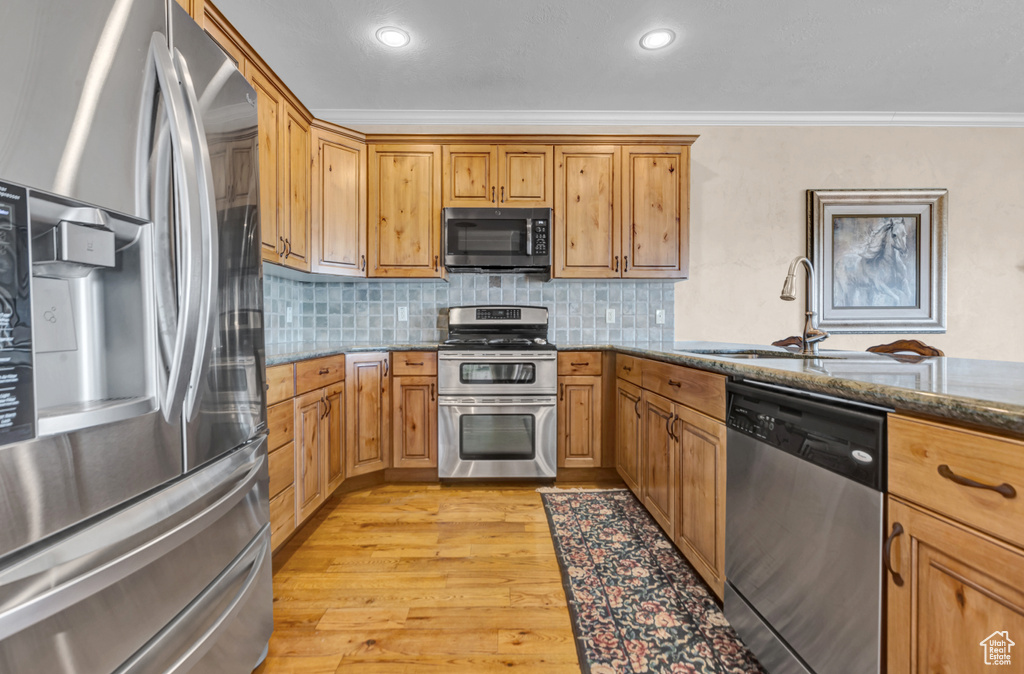 Kitchen with appliances with stainless steel finishes, crown molding, dark stone counters, light wood-style floors, and decorative backsplash