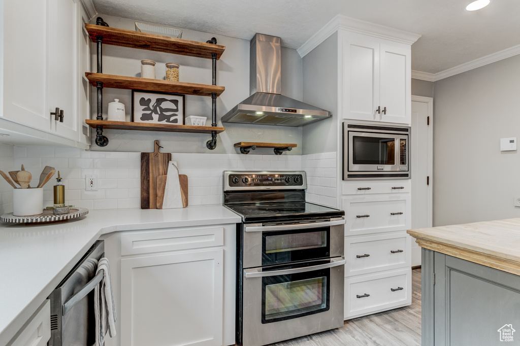 Kitchen with wall chimney exhaust hood, stainless steel appliances, light wood finished floors, ornamental molding, and tasteful backsplash