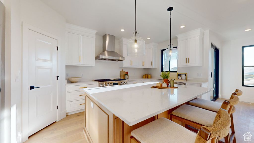 Kitchen with wall chimney exhaust hood, light wood finished floors, a kitchen bar, a kitchen island, and white cabinetry