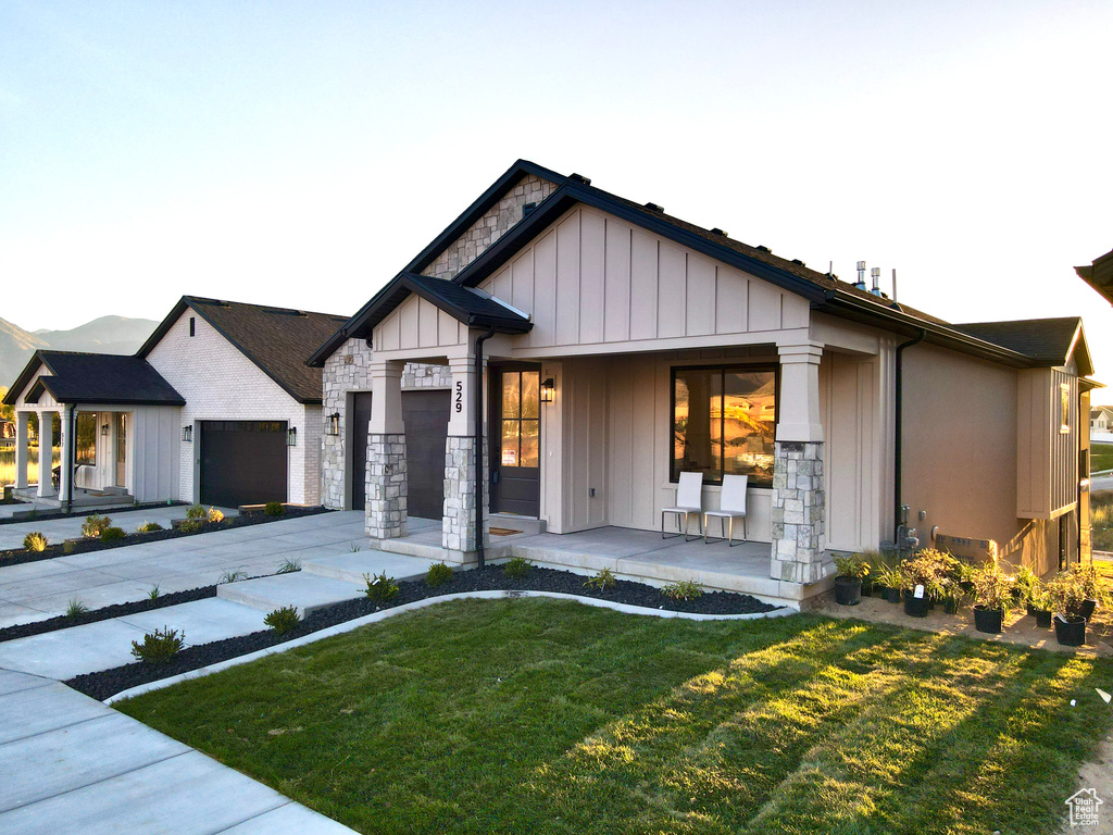 View of front of home featuring covered porch, a front lawn, stone siding, an attached garage, and board and batten siding