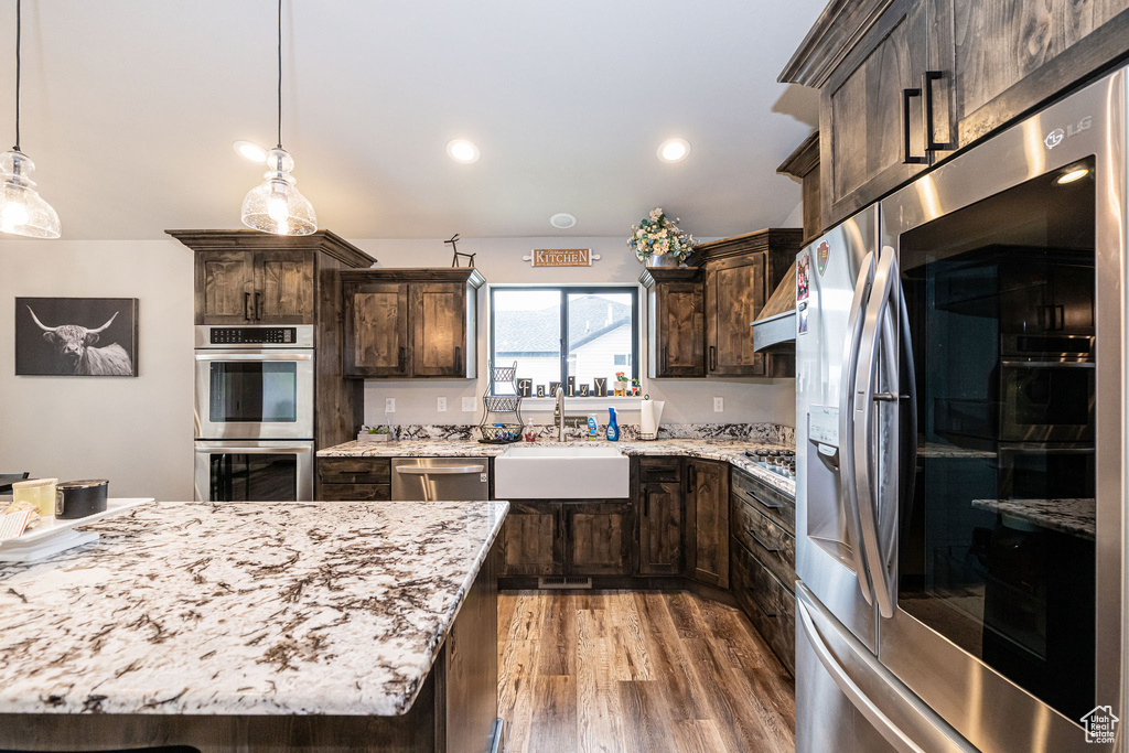 Kitchen featuring appliances with stainless steel finishes, dark brown cabinets, light wood finished floors, light stone counters, and hanging light fixtures