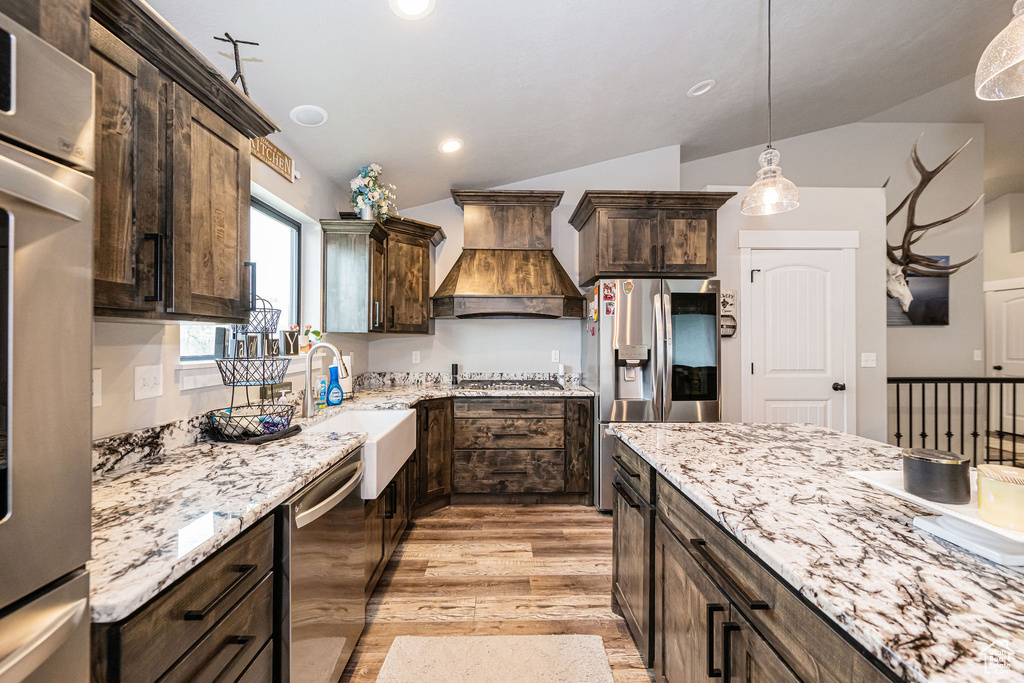 Kitchen featuring vaulted ceiling, appliances with stainless steel finishes, premium range hood, dark brown cabinets, and light wood finished floors