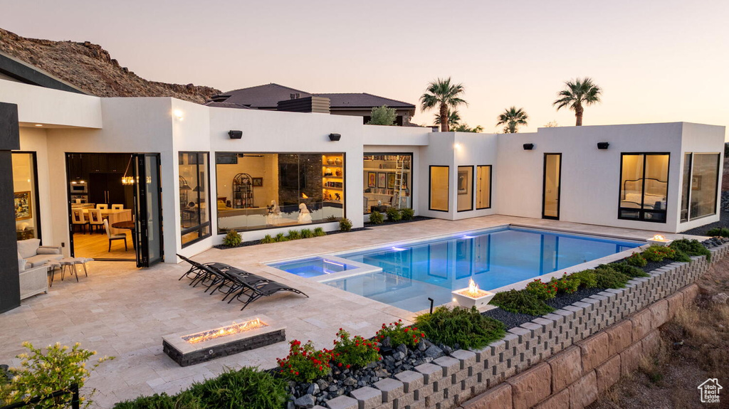 Back of house at dusk with an outdoor fire pit, a patio, stucco siding, and a pool with connected hot tub