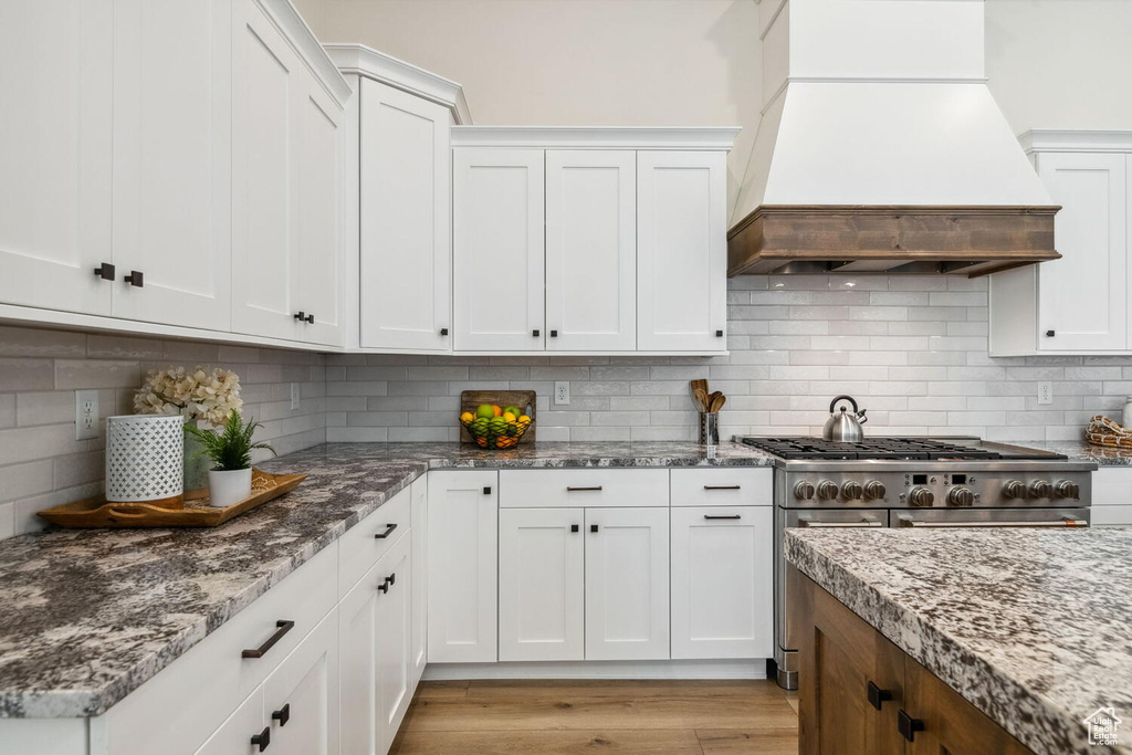 Kitchen featuring custom range hood, backsplash, white cabinetry, light stone counters, and light wood-style floors