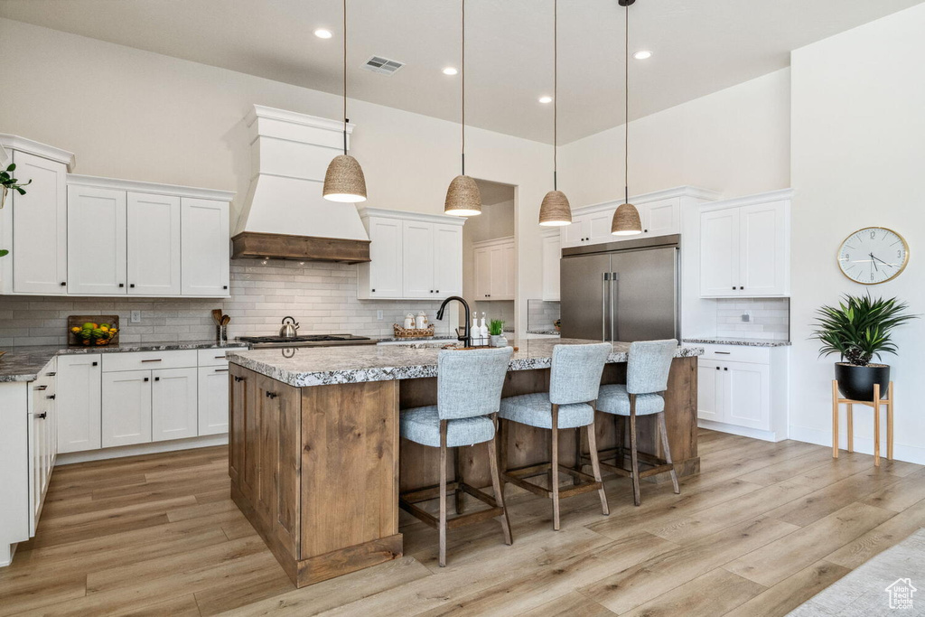Kitchen with wall chimney exhaust hood, stainless steel appliances, decorative backsplash, a kitchen island with sink, and light wood-style floors