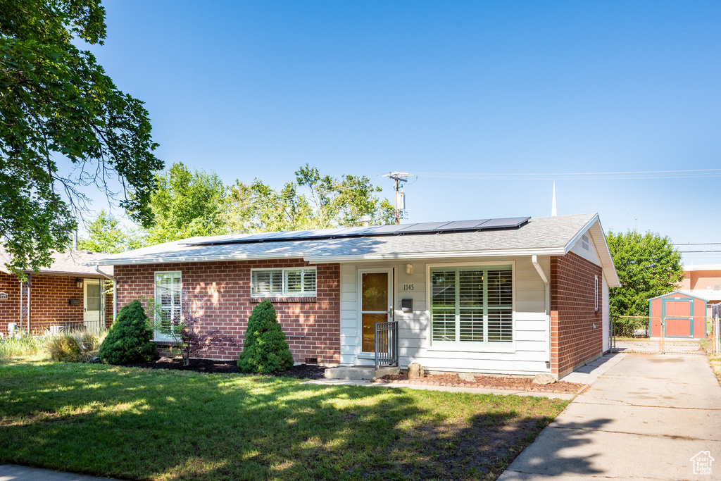 Single story home featuring a storage shed, a front lawn, roof mounted solar panels, and brick siding