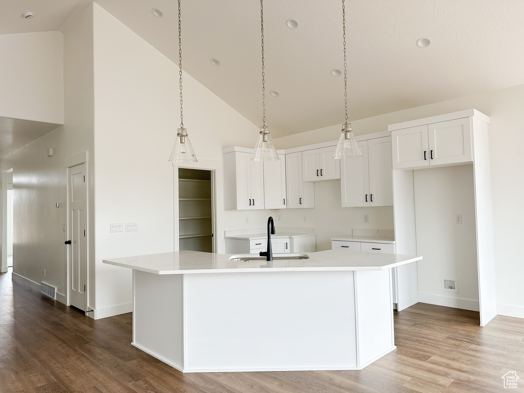 Kitchen featuring high vaulted ceiling, wood finished floors, white cabinetry, a center island with sink, and light countertops