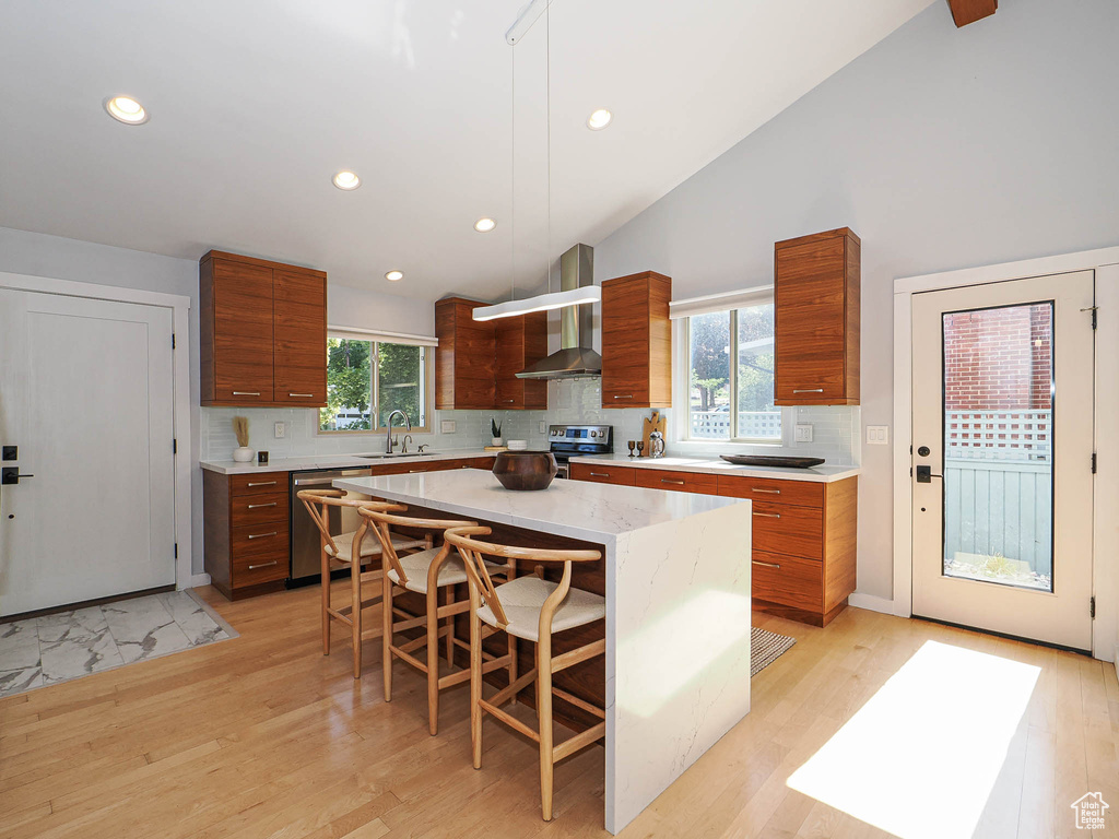Kitchen with stainless steel appliances, wall chimney exhaust hood, brown cabinetry, backsplash, and a kitchen bar