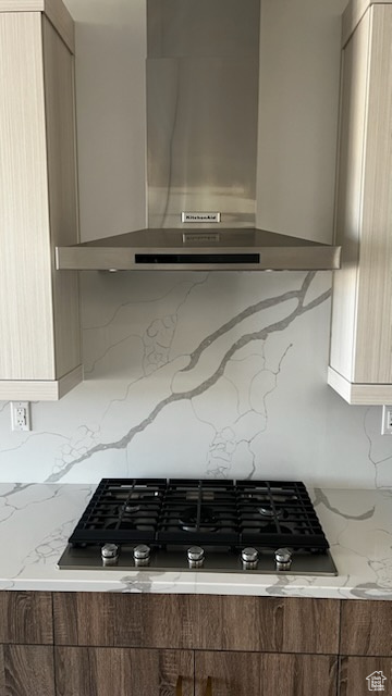 Kitchen with wall chimney range hood, backsplash, modern cabinets, black gas stovetop, and light stone counters