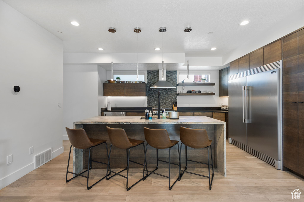 Kitchen featuring stainless steel built in fridge, wall chimney range hood, open shelves, a breakfast bar area, and recessed lighting